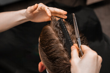 Stylist cuts male client hair using comb and scissors in barbershop closeup. Barber with...