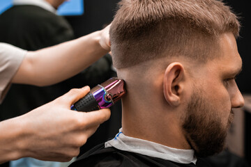Barber shaves client nape with electric shaver in barbershop closeup. Master does modern haircut to young man using cutting machine
