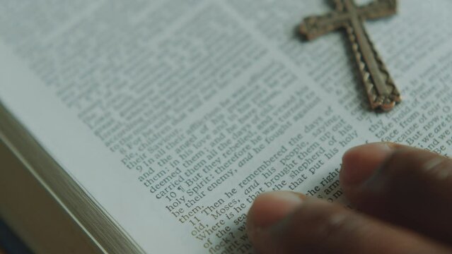 Hand of unrecognizable man reading religious text in Holy Bible with crucifix on it. Extreme close-up view