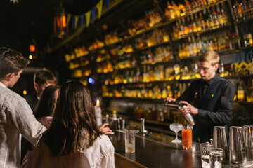 Group of guests sitting near bar counter supervises professional work of bartender. Young barkeeper prepares elite cocktails using metal shaker
