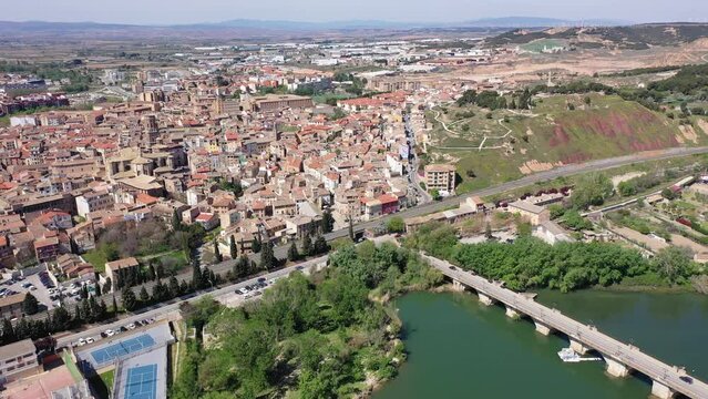 Picturesque aerial view of historic center of Tudela with brownish tiled roofs of residential buildings, medieval Cathedral of Saint Mary and ancient arched bridge over Ebro river on spring day, Spain
