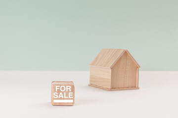 Simplistic wooden house model isolated on pale green background, with text For Sale on signboard.