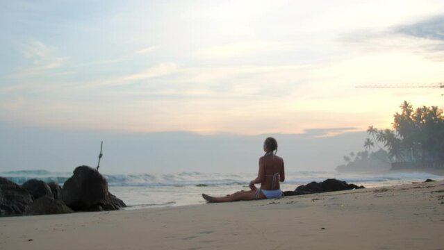 Woman silhouette practicing sitting straddle on sandy beach near ocean in mist. Lady athlete enjoys yoga session at summer sunset