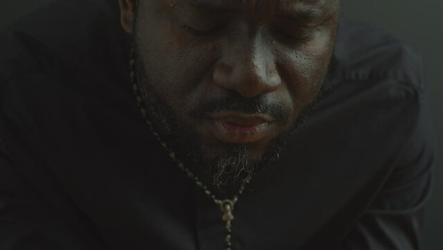 Black Catholic priest holding Holy Bible and saying a prayer with eyes closed. Close-up view, tilt-down shot
