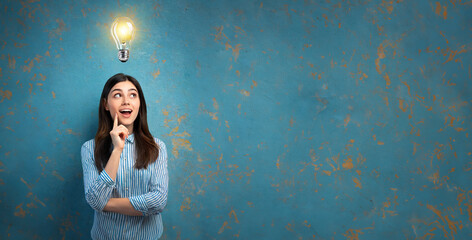 Illuminated light bulb above the head of a young woman, the concept of having an idea of a surprised girl with eureka on an old blue wall 