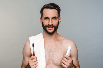 Healthy naked man with towel brushing his teeth isolated over grey background. Attractive young male model holding tooth brush and paste, doing morning daily routine