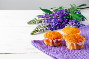 Soft fresh muffins on textile napkin. Delicious pastries and purple lupine flowers on white wooden background. Preparing dessert in confectionery. Copy space.