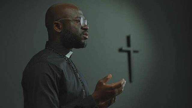 African American priest wearing cassock and rosary beads preaching a sermon during liturgical service in church. Side view, copy space