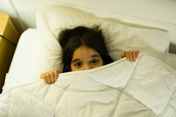 Hispanic little girl looking really scared at night and peeking out from under a white duvet in her bedroom