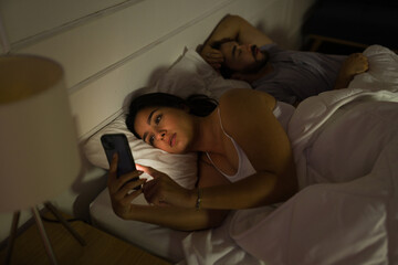 A woman lies awake, engrossed in her smartphone, while her partner sleeps beside her, depicting modern disconnect even at night