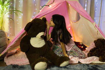 Hispanic young girl shares secrets with a big teddy bear in a magical bedroom tent beneath a starry night sky at home