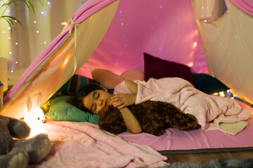 Hispanic child peacefully naps with her stuffed animal in a homemade tent, decorated with fairy lights at night