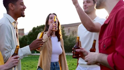 Group of young friends celebrating together cheering with beer bottles outside. Multiracial happy people laughing enjoying summer party outdoors. Community, youth and friendship concept.
