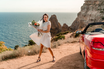 A woman in a white dress is standing next to a red convertible car. She is holding a bouquet of...