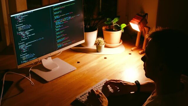 A young man programmer coding on a laptop in the dark with a view of the lights of the night city, color lighting in the room. Coding and hacking concept. Saint Petersburg, Russia - 20 April 2024