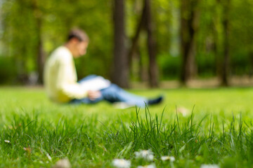 young man in blurred reading a book in a city park sitting on a green grass, outdoor recreation,...