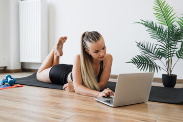 Young fit woman lies on yoga mat at home, browsing internet on laptop. Happy female with fitness...