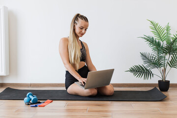 Young woman in sportswear sits on yoga mat in bright room, using laptop with fitness equipment nearby. Happy female looks engaged and content, highlighting blend of technology and healthy lifestyles.