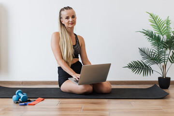 Fit young woman sitting on yoga mat in serene home environment, holding laptop. Caucasian female blends fitness routine with professional obligations, emphasizing lifestyle of health and productivity.
