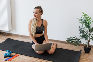 Young woman in sportswear sits on yoga mat in bright room, using laptop with fitness equipment nearby. Happy female looks engaged and content, highlighting blend of technology and healthy lifestyles.