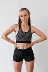 Young fit woman stands confidently against white background wearing fitness attire. Sportswoman in sports bra and black shorts, showcasing toned physique and focused expression.