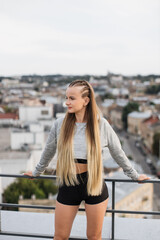 A young woman stands on a rooftop dressed in stylish athletic wear, overlooking a panoramic cityscape. Her relaxed posture and thoughtful expression reflect a moment of peace amidst urban life.