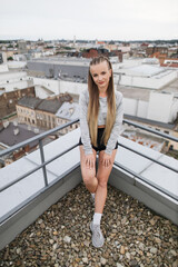 A young woman enjoys a moment of relaxation on a rooftop, overlooking an expansive cityscape. Dressed casually, she poses confidently, capturing the essence of urban living and youthful freedom.