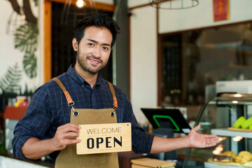 Smiling male coffee shop staff holding 'Open' sign, casual yet stylish work apron, inviting...
