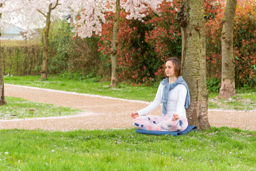 A young woman meditates in the lotus position, leaning against a tree trunk against the backdrop of spring cherry blossoms. Concept of mental and physical health and balance in life.