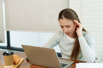 Beautiful young Asian woman Coffee shop owner sitting in front of a work desk in front of a laptop computer. Check sales work from notebook Hands gently massage the head showing a stressed expression