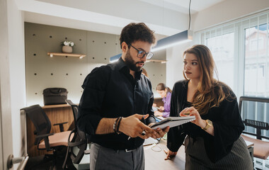 A male and female business professional engage intensely over a digital tablet in a well-lit office space, indicating collaboration and strategic planning.