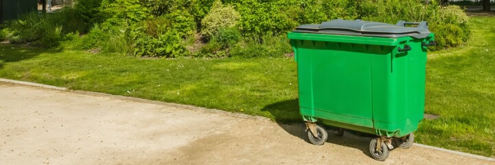 Large green waste container on wheels placed beside a footpath in a public park, symbolizing urban cleanliness and environmental awareness, related to Earth Day
