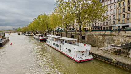 Cloudy day over the Seine River in Paris, with a moored riverboat and early spring foliage lining the historic stone quays, ideal for travel and urban themes