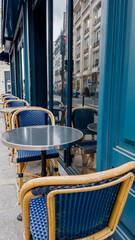 Empty blue wicker chairs at a Parisian sidewalk cafe with a reflective metal table, evoking...