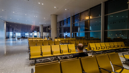 Lone traveler sitting in an empty airport waiting area with yellow seats, symbolizing travel,...