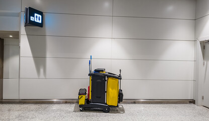 Empty airport cleaning cart with bright yellow detailing parked beside restrooms sign, depicting...