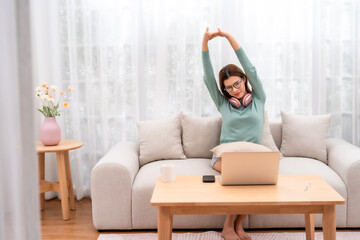 A serene woman enjoys music on her headphones while lounging on a cozy sofa, stretching and...