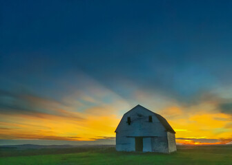 Old white barn on the edge of a farm field silhouetted by the sunset