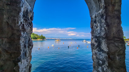 A view through a stony arch on the cities located along the coastal line of Istria, Croatia. There are plenty of buoys in the water and a few boats parked in there. The Mediterranean Sea is calm.
