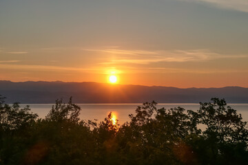 The sun setting behind mountains in Croatia. The Mediterranean Sea is calm and clear, reflecting the sunbeams. The sky is orange and covered with clouds. Dense tree crowns on the bottom. Remedy