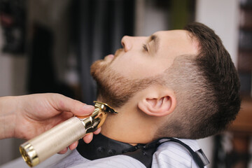 Professional hairdresser uses a hair clipper for fringing beard for a handsome man in barbershop....