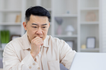 Fototapeta na wymiar Mature Asian businessman in thoughtful pose, sitting at his office desk. This image captures a moment of deep concentration and decision-making in a professional setting.