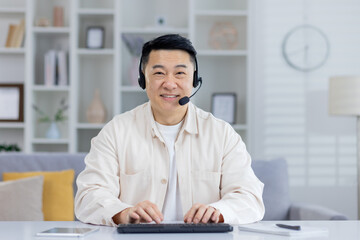 A middle-aged Asian man smiles warmly while using a headset and working on a keyboard in a bright,...