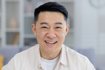 Obraz premium Portrait of a cheerful middle-aged Asian man with a radiant smile, wearing a light shirt, seated in a bright, modern living room.
