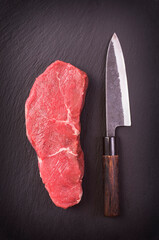 Raw dry aged angus roast beef steak offered as top view  on a design board with a Japanese santoku knife