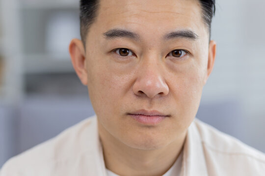 A detailed close-up portrays a middle-aged Asian man with a contemplative, serious expression, captured indoors with a soft focus background.