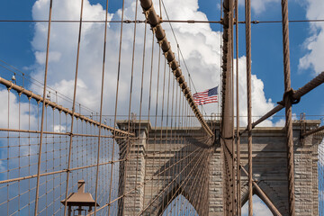 The gate of Brooklyn Bridge with a waving American flag on top of it contrasted with a blue sky with puffy, white clouds. Suspension bridge in New York City. Massive construction. Local landmark