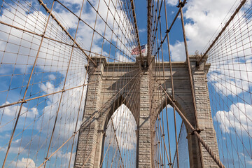 The gate of Brooklyn Bridge with a waving American flag on top of it contrasted with a blue sky with puffy, white clouds. Suspension bridge in New York City. Massive construction. Local landmark
