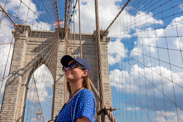 Woman in sunglasses and a cap standing in from of a gate of Brooklyn Bridge with a waving American...