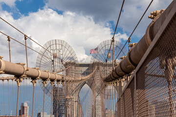 The gate of Brooklyn Bridge with a waving American flag on top of it contrasted with a blue sky with puffy, white clouds. Suspension bridge in New York City. Massive construction. Local landmark - Powered by Adobe